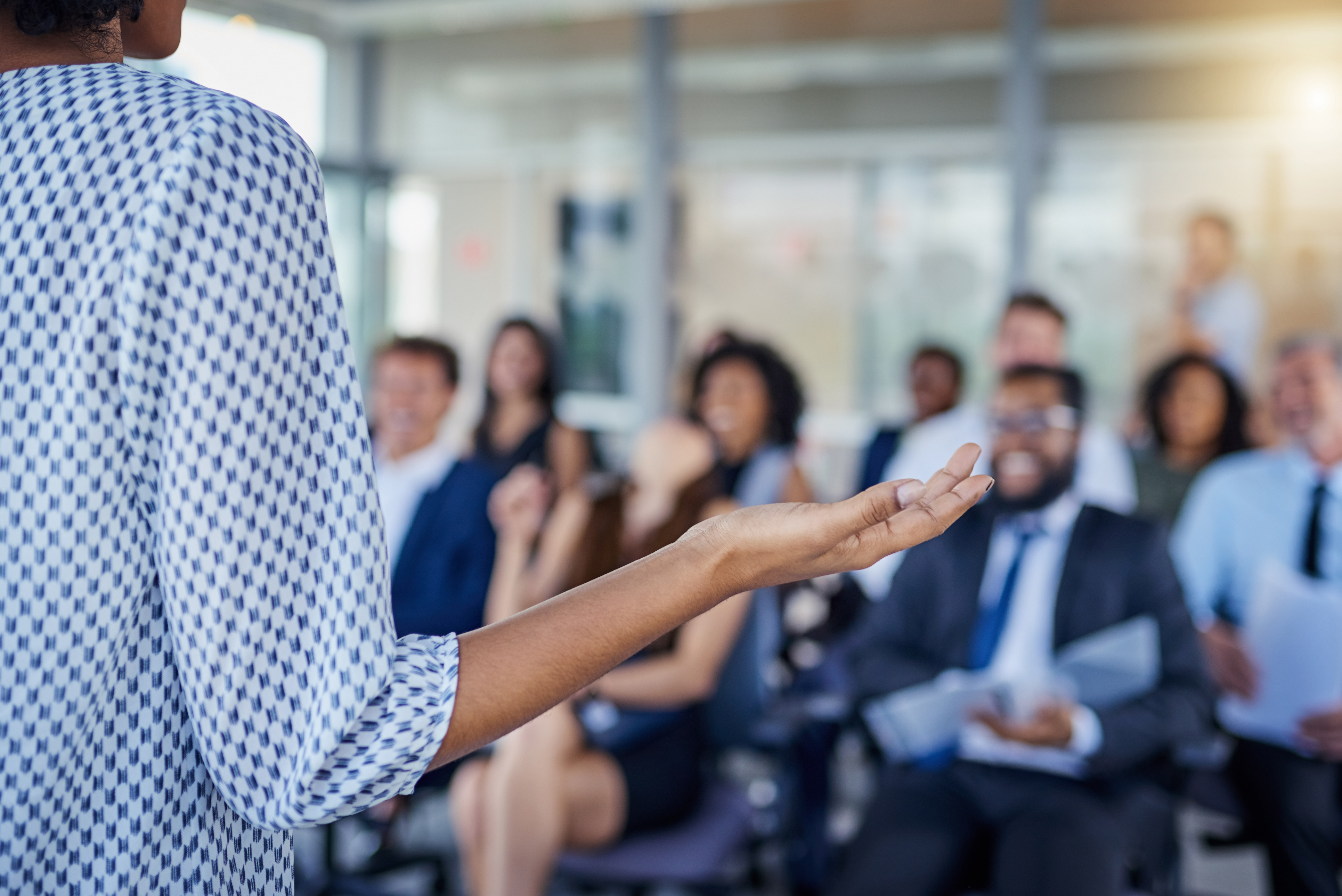 Woman addressing room full of individuals dressed in business attire.
